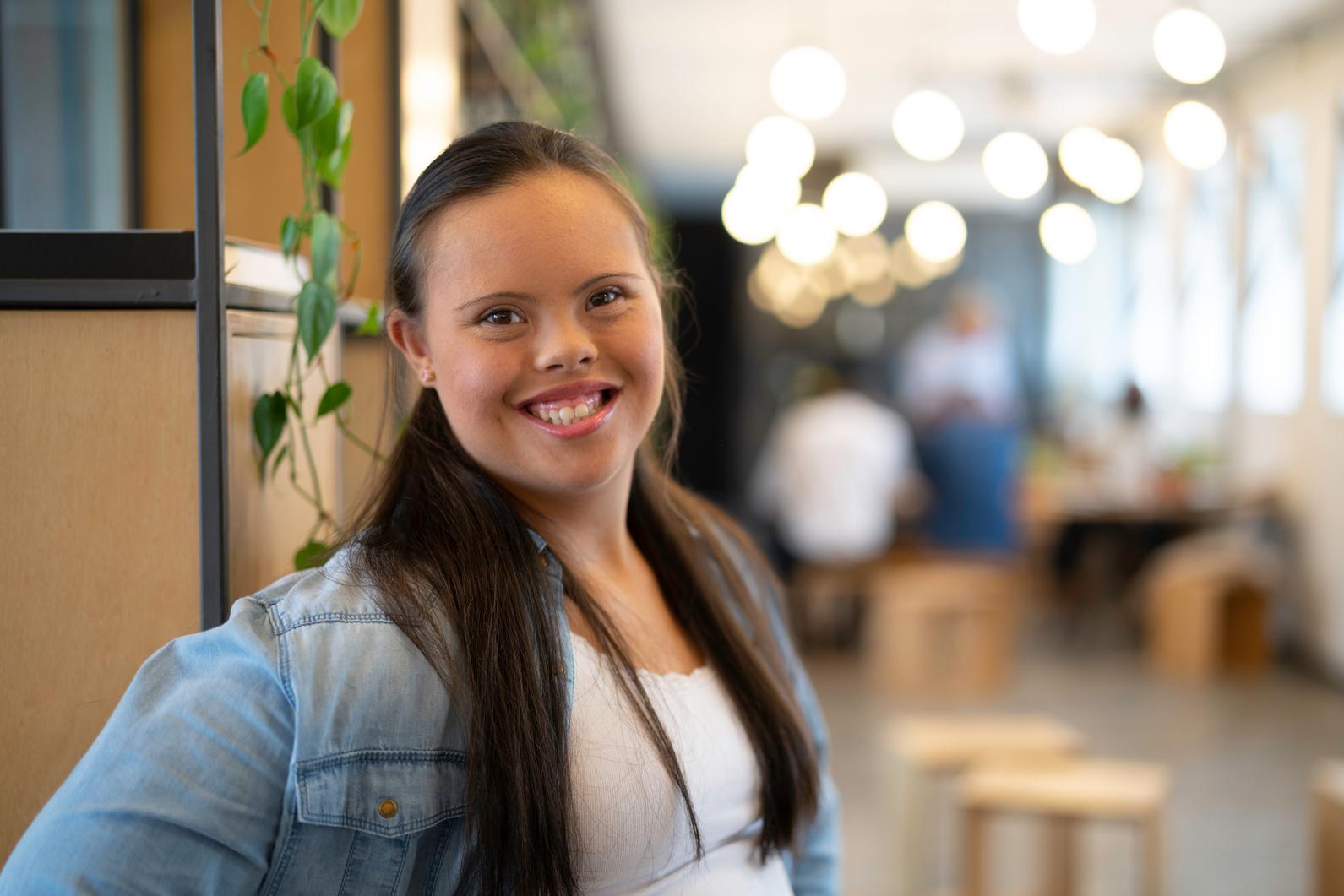 A young woman with Down syndrome smiling warmly, wearing a denim shirt in a bright café setting